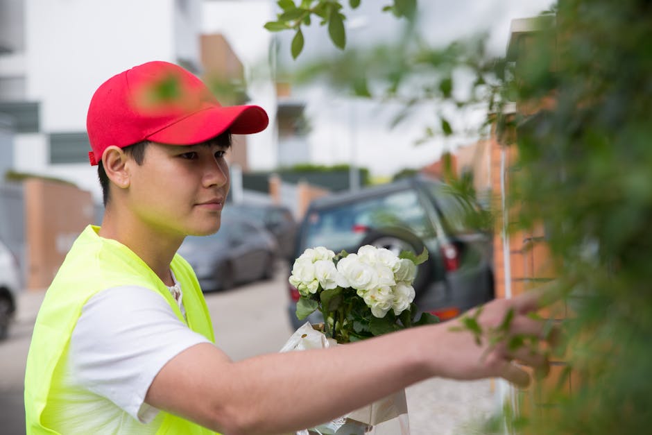 A delivery person wearing a red cap and a yellow high-visibility vest is holding a bouquet of fresh white hydrangeas wrapped in white paper. They are standing outdoors on a residential street, reachin