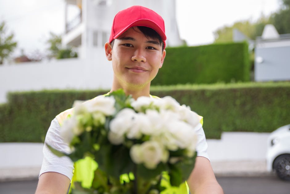 A delivery person wearing a bright yellow high-visibility vest and a red cap is standing outdoors, smiling warmly, and holding a bouquet of fresh white flowers with green foliage. The bouquet appears 