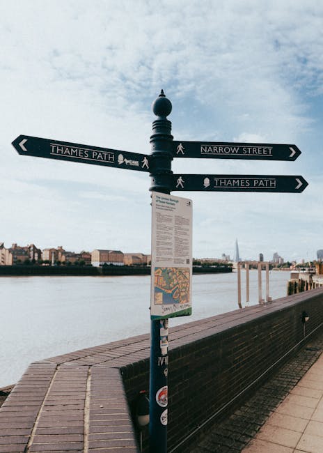 The image features a black, decorative street sign pole situated along the Thames Riverside, with directional signs pointing towards Thames Path and Narrow Street. The sign is mounted on a brick-paved