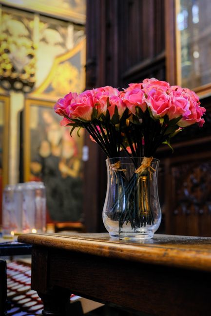 A clear glass vase filled with a bouquet of fresh pink peonies with lush, ruffled petals and dark green stems, placed on a rustic wooden table. The flowers are arranged in a loose, natural style, show