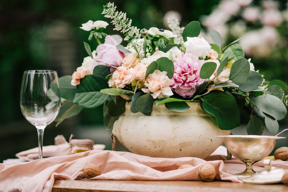 A large, elegant floral arrangement featuring pink peonies, light peach carnations, and white astilbe with green eucalyptus leaves, displayed in a rustic, off-white ceramic bowl. The bouquet is set on
