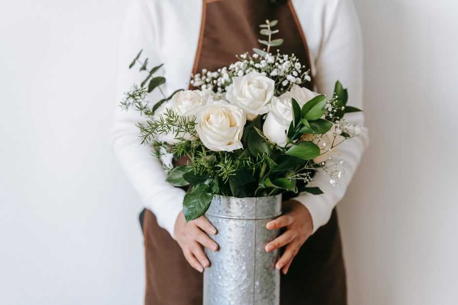 A person holding a tall, cylindrical silver vase filled with an elegant floral arrangement. The bouquet features large, creamy white roses with delicate, tightly packed petals, complemented by sprigs